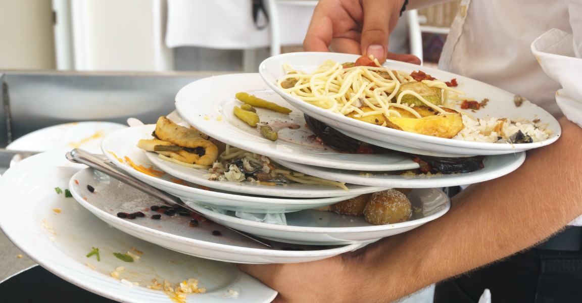 A male server carrying eight dirty plates with food scraps on them to toss in the trash.