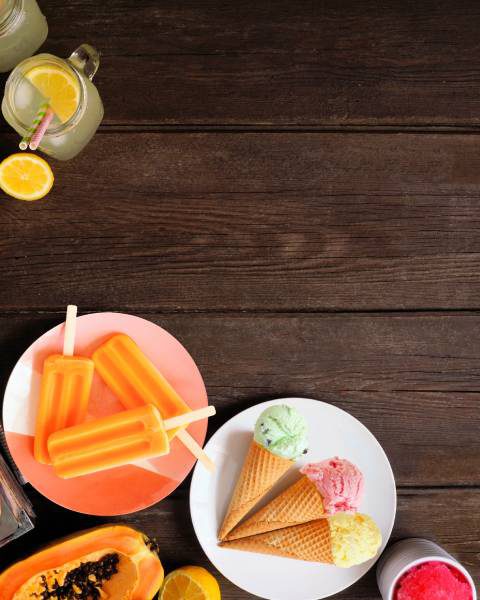 A wooden table with colorful treats, including plates of red and orange popsicles, ice cream cones, and tubs of ice cream.