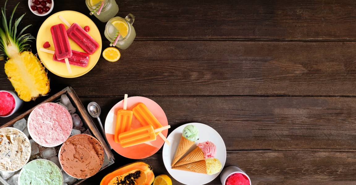 A wooden table with colorful treats, including plates of red and orange popsicles, ice cream cones, and tubs of ice cream.
