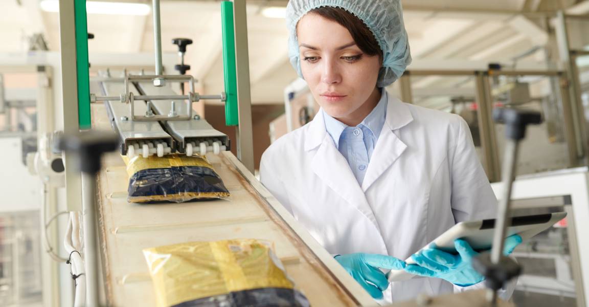 A worker at a food processing plant inspects sealed bags as they climb up a conveyor belt.