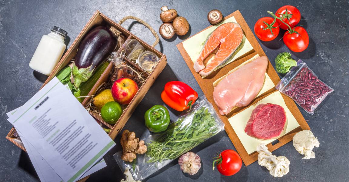 The ingredients of a meal delivery kit displayed on a black surface. Some recipe papers are lying on top of the box.