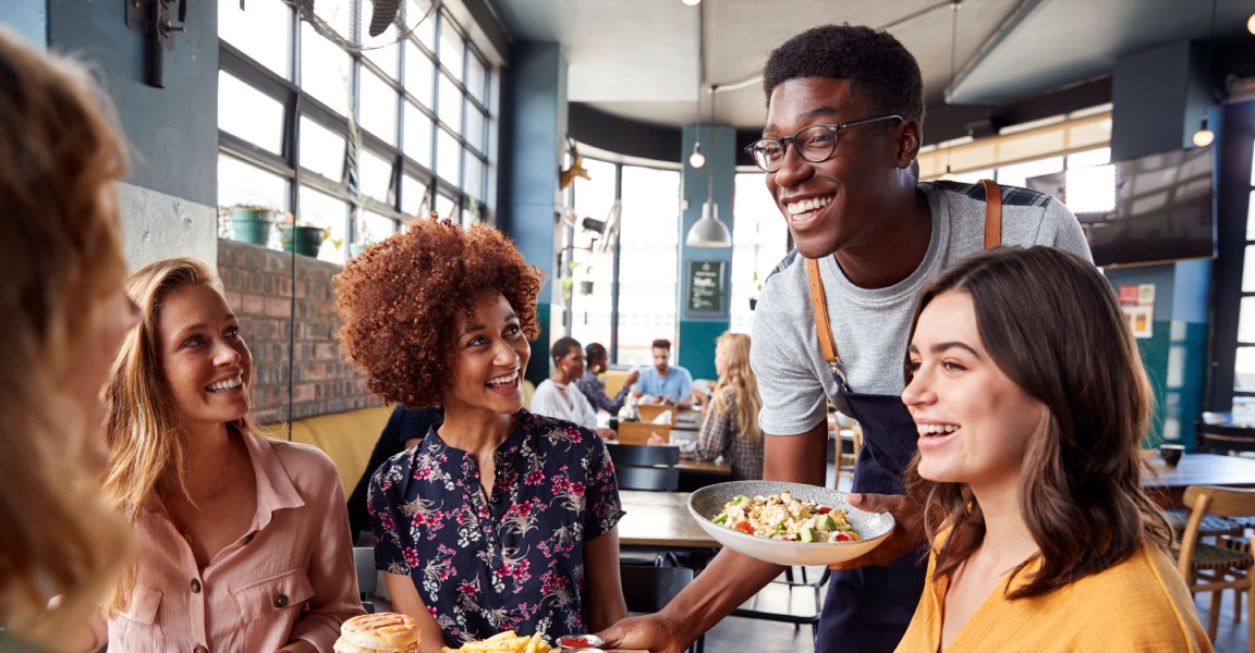 A group of young women sitting down for a meal at a restaurant. A smiling, friendly-looking waiter serves them their food.